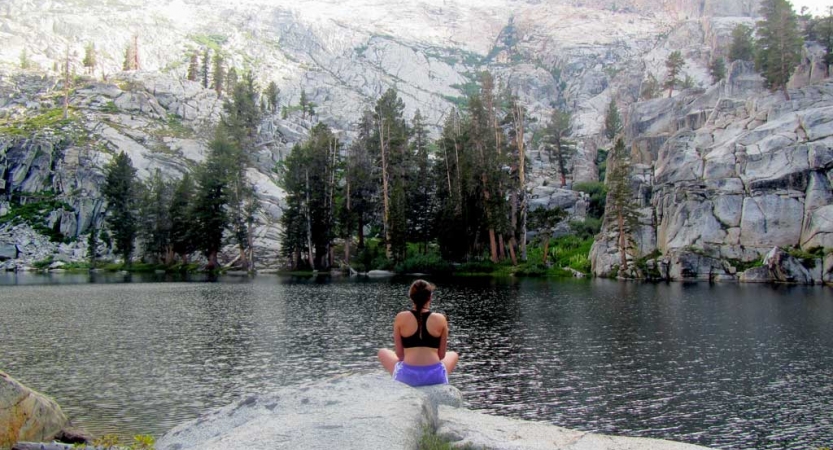 A person sits on the shore of a body of water, facing away from the camera. The water is framed by a steep rocky landscape. 